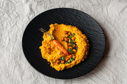 M&ouml;hren Hummus auf dunklem Teller mit Kichererbsen und ger&ouml;steter M&ouml;hre als Garnitur Leinentuch im Hintergrund minimal