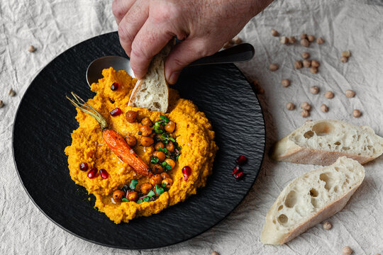M&ouml;hren Hummus auf dunklem Teller mit Kichererbsen und ger&ouml;steter M&ouml;hre als Garnitur Leinentuch im Hintergrund und eine Hand die mit einem St&uuml;ck Baguette etwas nimmt