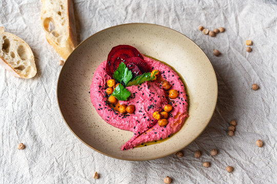 Rote Bete Hummus auf hellem texturierten Teller mit Kichererbsen Garnitur Leinentuch im Hintergrund