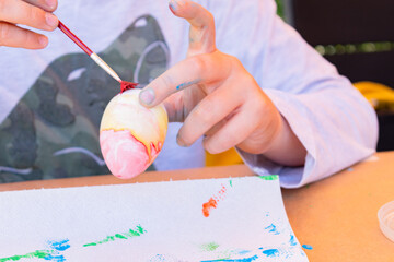 11-year-old boy watercolour painting a yellow and red Easter egg with a paint brush on a table in his backyard.