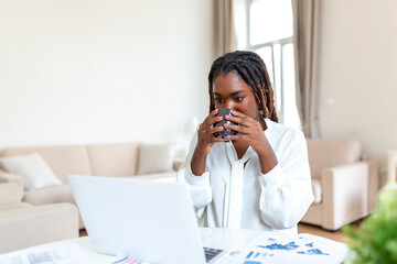 Young African American businesswoman working remotely from home using a laptop and enjoying a cup...