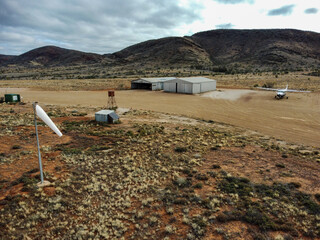 Aeroplane by old hangars and windsock at gravel airstrip with mountains behind