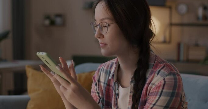 Slow motion of cheerful girl student using smartphone messaging in social media smiling in apartment in evening. Gadget and communication concept.