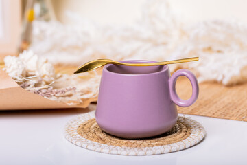 Amidst an array of eclectic boho decorations, a charming scene unfolds on a pristine white table: a sleek purple coffee mug, emanating warmth, paired with a delicate golden spoon.