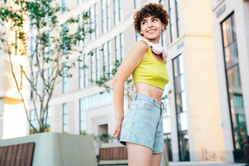 Young beautiful smiling hipster woman in trendy summer clothes. Carefree woman with curls hairstyle, posing in the street at sunny day. Positive model outdoors. Listens music at her headphones on neck