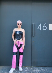 Young gay boy with pink hair and make-up and dressed in pink and black is leaning against a black...