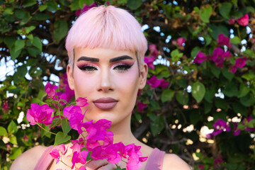 Portrait of young gay boy with pink hair and make-up has next to his face a purple bougainvillea...