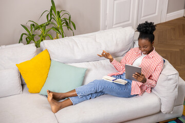 Creative woman with afro hairstyle making notes in diary while working on tablet.