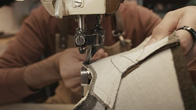 Zoom in shot of shoemaker sitting behind sewing machine in workshop and stitching leather material