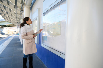 Fototapeta premium Full length side view of a female traveler standing at a board with train schedule information