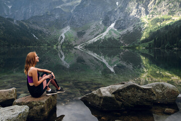 Young blonde fit and sporty woman in sportswear sitting on the rock on the shore and admiring beautiful view of green hills and mountains on Morskie Oko lake, High Tatras, Zakopane, Poland. - obrazy, fototapety, plakaty