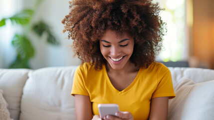 young woman with curly hair, wearing a yellow shirt, smiling and looking down at her smartphone - obrazy, fototapety, plakaty