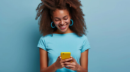 smiling young woman looking down at her yellow smartphone with pleasure, wearing a blue top and matching blue hoop earrings against a soft blue background - obrazy, fototapety, plakaty