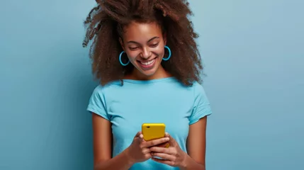 Foto op Plexiglas smiling young woman looking down at her yellow smartphone with pleasure, wearing a blue top and matching blue hoop earrings against a soft blue background © MP Studio