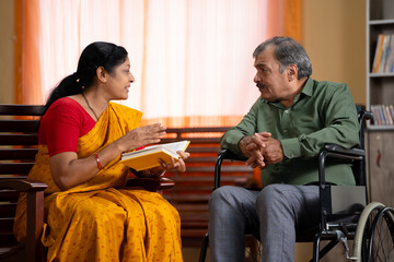 caring Indian senior wife telling by reading novel or story from book to husband sitting on...