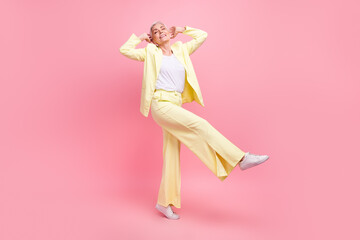 Full size photo of good mood woman with short hairdo wear yellow suit hold hands behind head dancing isolated on pink color background