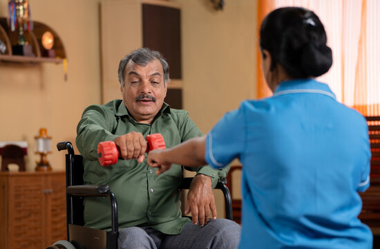 Shoulder shot Indian nurse encouraging to lift dumbbell during to elderly man on wheelchair with disability at home during physiotherapy session - concept of medical counselling and rehabilitation.