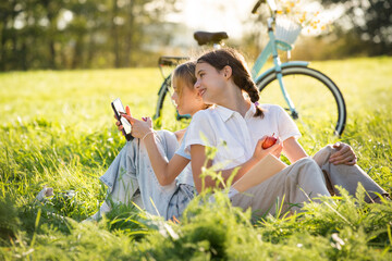 Two girls spend time on green grass lawn in park together, scroll through smartphones, read books,...