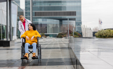 A young beauty in a wheelchair is taken by a nurse for a walk on the street near the hospital