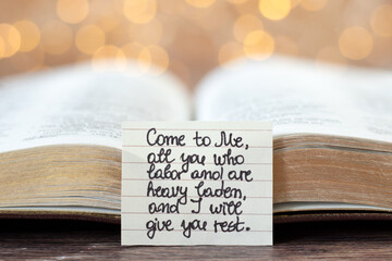 Handwritten quote for Christians to come to God Jesus Christ's rest in front of an open holy bible book with bokeh light background. Close-up. Selective focus. Biblical concept.