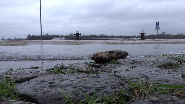 Low angle view of flooded road and quay in Tolkamer, with cargo vessels and ships in the background. High water levels of the Rhine river and flood risk in the Netherlands and Northwestern Europe.
