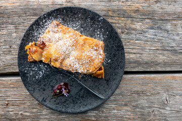 Delicious crispy oven-fresh apple strudel dessert lovingly served on a black marbled plate decorated with wild flowers and jam arranged on a wooden table