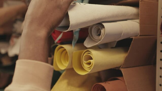 Close up shot of hands of male cobbler touching rolled suede leather of white and yellow colors while choosing material for manufacturing shoes in workshop