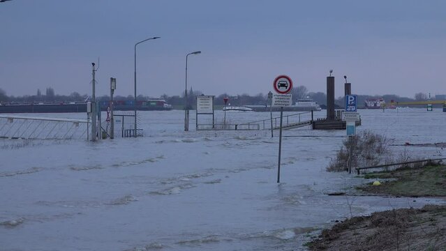 Overflown road and quay in Tolkamer, with cargo vessels and boats in the background. High water of the Rhine river in the Netherlands and Northwestern Europe.