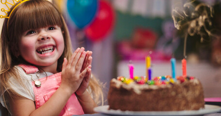 Close up of cheerful little cute girl in birthday crown celebrating her birthday, blow colorful candles on the cake and smiling.