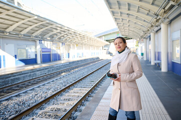 Attractive young woman commuter in beige coat, waiting to board the train on the railway station, standing on the platform with a smart phone in hands. People. Railroad trip