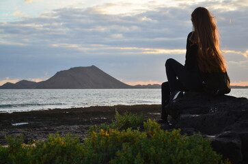 Young woman with long brown hair sitting on the volcanic beach of Playa de la Barreta on the island...
