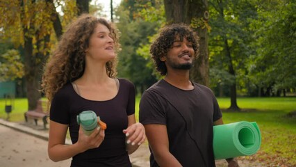 Happy young girlfriend boyfriend walk street together multiracial Caucasian woman Indian man talking laughing going on fitness training in city carrying water bottle and fit mat sport yoga outdoors - Powered by Adobe