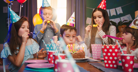 Caucasian small cute birthday boy blowing the candles on he cake while his happy friends in...