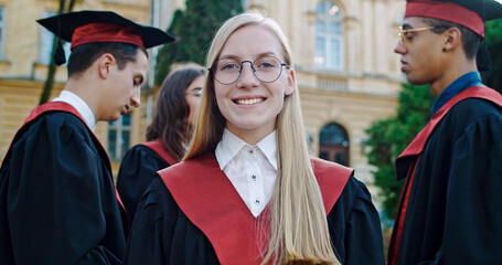 Caucasian blond pretty young female master graduate smiling and looking straight to the camera, then taking off glasses. Graduation ceremony. Portrait.