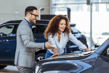 Happy woman with car dealer in auto show or salon. Car Sales Manager Showing Auto To Lady Buyer. Beautiful caucasian female client customer choosing new car.