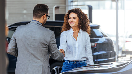 Happy salesman congratulating his female customer for buying a new car in a showroom. Woman buying...