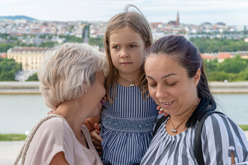 An elderly woman of 60-70 years old and a woman of 40-45 years old with her daughter stand next to each other on a blurred background of the urban landscape, an emotional family meeting of different 