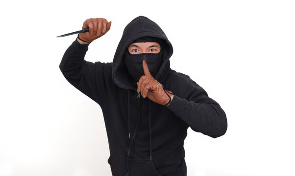 A man wearing a black hoodie, balaclava and gloves raised a knife while the other finger on his lips; threatening expression