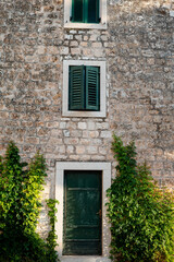 Fototapeta na wymiar Green door of ancient cottage in stone wall decorated with climbing plants. Fresh herbs growing upwards creating cozy atmosphere in city Supetar, Croatia. Concept of comfort and buildings.