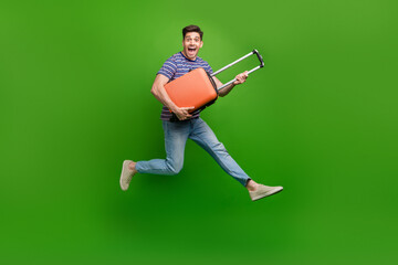 Full length photo of ecstatic man wear stylish t-shirt jeans trousers jumping with baggage in hands isolated on green color background