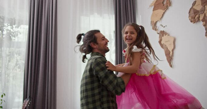 Happy brunette man in a checkered green shirt picks up and spins his little daughter in a pink fairy costume in his modern home