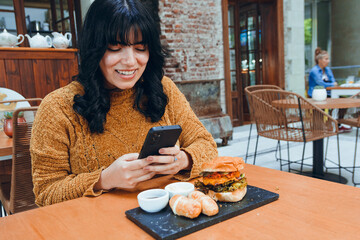 young Latin woman posting with her phone on social networks a photo of hamburger she is eating
