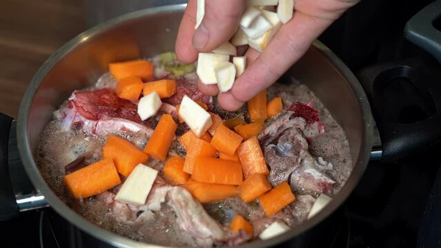 Person adding chopped parsnips to bone broth beginning to boil in pot.