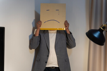 Unhappy man employee with box instead of his head. Portrait of businessman covering his face with paper mask with sad face drawn on it. - obrazy, fototapety, plakaty