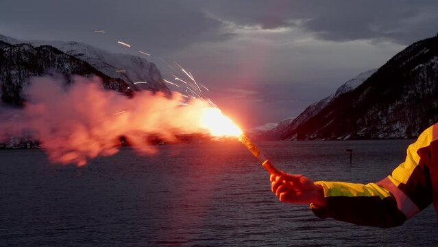 Ship crewman holds distress signal flare, snowy mountains, fjord in background.