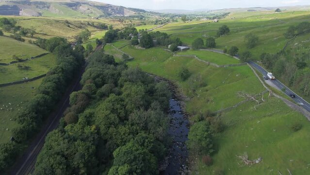 Drone footage flying over a beautiful shallow tree and grass lined river valley in sunny rural Yorkshire, UK. The valley is bordered by a country road and railway with hills in the distance