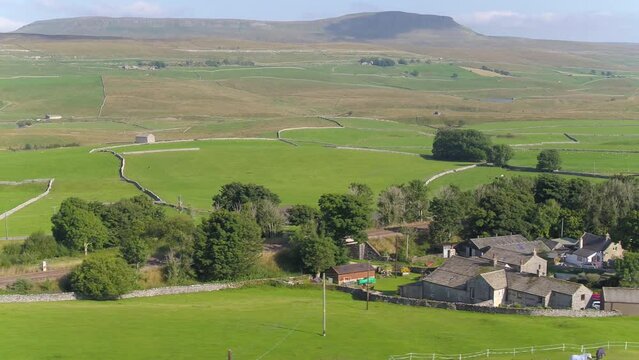 Drone footage on a sunny summer day in the village of Selside, North Yorkshire moving sideways over farmland and fields, dry stone walls with Pen Y Ghent mountain in the distance