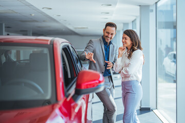 Beautiful young couple at car showroom choosing a new car to buy. Happy beautiful couple is choosing a new car at dealership. Happy young couple standing alongside their dream car and looking in