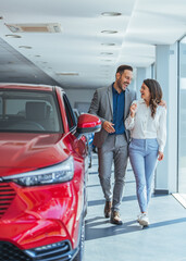 Beautiful young couple at car showroom choosing a new car to buy. Happy beautiful couple is choosing a new car at dealership. Happy young couple standing alongside their dream car and looking in