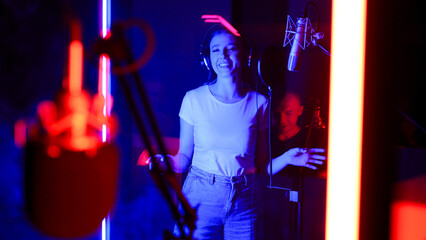 Young woman behind the glass of a recording studio with headphones records a song into a microphone.